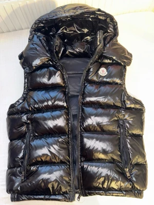 Moncler väst med huva - Svart glansig moncler väst. Inga skador eller liknande. Den är i perfekt skick. Storlek S. Västen är äkta och darför är den rätt så dyr pris kan diskuteras!
