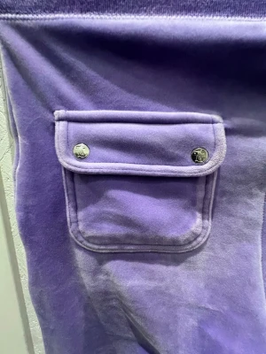 Lila mjukisbyxor Juicy Couture XS - Säljer ett par lila mjukisbyxor från Juicy Couture i mjuk velour. Byxorna har en ficka med lock och tryckknappar på sidan, samt bred resår i midjan. Perfekta för en chill och trendig look. Ny pris är 1300kr. 