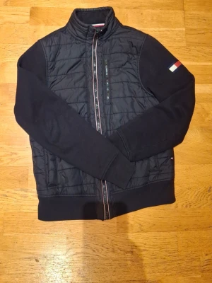 Svart quiltad cardigan från Tommy Hilfiger st xs - Svart cardigan från Tommy Hilfiger med quiltad front och ärmar i sweatshirtmaterial. Hög krage, dragkedja framtill och en liten flaggdetalj på ärmen. Sportig och stilren design som funkar till det mesta.