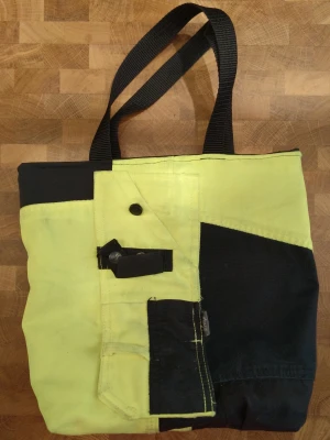 Gul och svart shoppingväska med fickor - Unik shoppingväska i gul och svart canvas med flera praktiska fickor och detaljer. Väskan har svarta handtag i slitstarkt material och coola kontrastsömmar. Perfekt för dig som vill sticka ut och gillar funktionella accessoarer med arbetsklädesvibbar. Väskan är vändbar. Sydd av gamla arbetsbyxor. Det svarta canvas tyget är lite slitet med några små hål men inget som stör funktionen.
