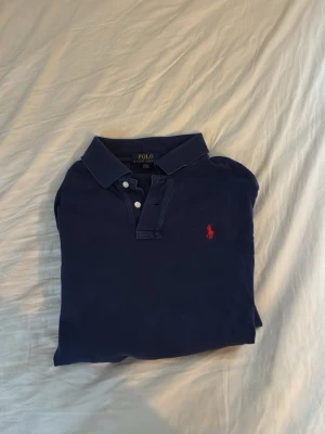 Marinblå långärmad piké från Polo Ralph Lauren - Klassisk marinblå långärmad pikétröja från Polo Ralph Lauren med röd broderad logga på bröstet. Tröjan har krage, knappar vid halsen och är tillverkad i mjuk bomull. Perfekt för en stilren och avslappnad look.är långarmmad