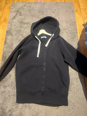 Svart zip hoodie med vita snören - Snygg svart hoodie med dragkedja och vita snören i huvan. Klassisk känguruficka framtill och ribbade muddar vid ärmar och nederkant. Perfekt för en avslappnad streetwear-look.