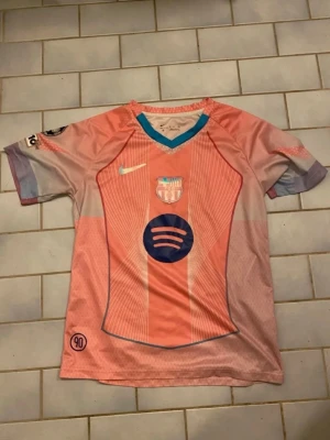 Rosa FC Barcelona matchtröja Nike - Snygg rosa FC Barcelona matchtröja från Nike med klubbmärke och sponsorlogga på bröstet. Tröjan har korta ärmar, V-ringning med turkos kant och detaljer i lila och blått. Perfekt för dig som älskar fotboll och vill sticka ut på planen.