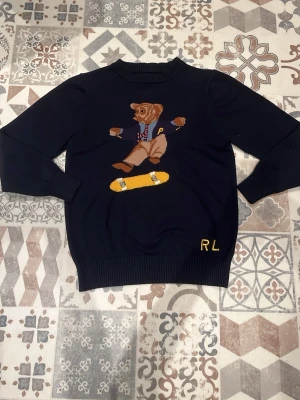 Mörkblå stickad tröja med björnmotiv - Snygg mörkblå stickad tröja från Ralph Lauren med ett stort broderat motiv av en björn på skateboard på framsidan. Tröjan har rund hals, ribbade muddar och RL-broderi i gult nedtill. Perfekt för dig som gillar unika och lekfulla plagg.