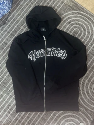 Svart zip hoodie från Hoodrich - Snygg svart hoodie med dragkedja från Hoodrich. Stor vit logga broderad över bröstet och klassisk huva. Tillverkad i mjukt material, perfekt för en avslappnad streetwear-look. Fickor framtill och ribbade muddar vid ärmar och nederkant.