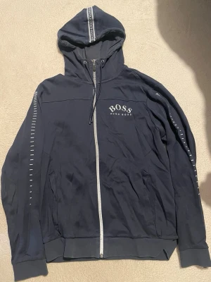 Marinblå hoodie från Hugo Boss - Snygg marinblå hoodie från Hugo Boss med dragkedja framtill och vit logga på bröstet. Huva med dragsko och dekorativt band längs huvan. Vita detaljer längs ärmarna ger en sportig look. Perfekt för en avslappnad stil.