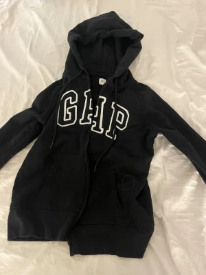 Svart hoodie med dragkedja från GAP - Säljer en svart hoodie från GAP med stor vit logga på bröstet. Hoodien har dragkedja, kängurufickor och justerbar huva med snören. Perfekt för en avslappnad och cool stil. Materialet är mjukt och bekvämt, troligen bomullsblandning.