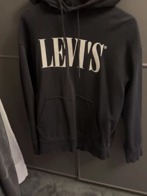 Svart hoodie från Levi's med logga - Svart hoodie från Levi's med stor vit logga framtill. Klassisk känguruficka och justerbar huva med snören. Tillverkad i mjukt bomullsmaterial som känns skönt mot huden. Perfekt för en avslappnad och trendig look.