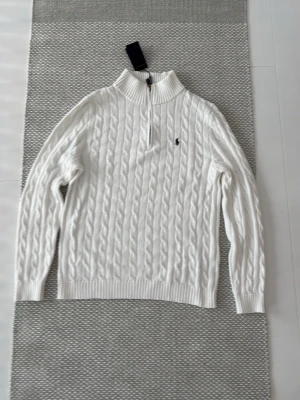Vit kabelstickad tröja från Polo Ralph Lauren - Snygg vit kabelstickad tröja från Polo Ralph Lauren med halv dragkedja och hög krage. Tröjan har ett broderat svart logotyp på bröstet och ribbade muddar. Perfekt för dig som gillar klassisk och stilren design.