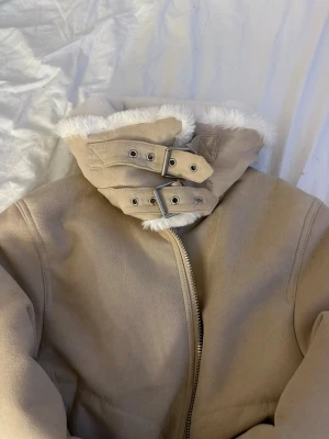 Beige teddyfodrad kappa med spännen - Snygg beige kappa med mjukt vitt teddyfoder och två dekorativa spännen vid kragen. Dragkedja framtill och hög krage som skyddar mot kylan. Perfekt för dig som vill ha en trendig och varm look under höst och vinter.