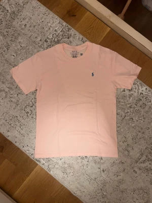 Ljusrosa t-shirt från Polo Ralph Lauren - Säljer en ljusrosa t-shirt från Polo Ralph Lauren med den klassiska blå broderade loggan på bröstet. T-shirten har rund hals och korta ärmar, perfekt för en clean och stilren look.