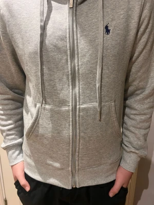 Grå zip hoodie från Ralph Lauren - Snygg gråmelerad hoodie från Ralph Lauren med dragkedja framtill, två stora fickor och justerbar huva med snören. Klassisk blå logga broderad på bröstet. Tillverkad i mjukt sweatshirtmaterial som är riktigt skönt att ha på sig.