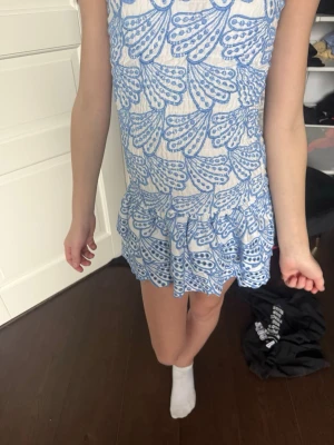 Blåvit kortklänning med volang - Söt kortklänning med blått broderat mönster på vit botten. Klänningen har smala axelband, volangkant nedtill och en figurnära passform. Perfekt för sommarens varma dagar och har en fräsch, ungdomlig stil.