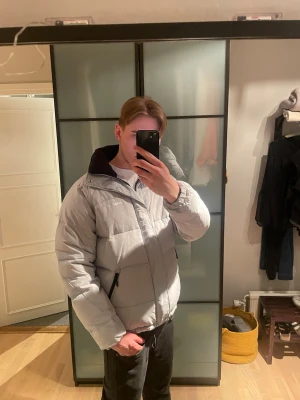 Ljusgrå pufferjacka från Zara - Säljer en stilren ljusgrå pufferjacka från Zara med svart foder och dragkedja framtill. Jackan har två sidofickor med dragkedja och hög krage för extra värme. Perfekt för kalla vinterdagar och ger en clean look.