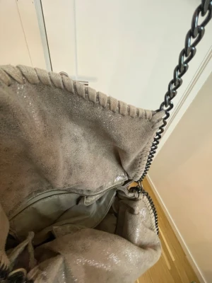 Grå axelremsväska med kedjedetaljer - Säljer en snygg grå brun handväska med glittrig yta och kraftiga kedjor som axelrem. Väskan har dekorativa sömmar längs kanten och en rymlig insida med innerfack. Materialet ser ut som mockaimitation och kedjorna är i mörk metall.