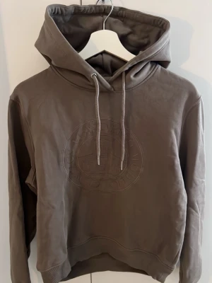 Brun hoodie från Bondelid - Snygg brun hoodie från Bondelid med broderad logga och motiv på bröstet. Hoodien har en stor huva med dragsko och ribbade muddar vid ärmslut och nederkant. Perfekt för dig som gillar klassisk och stilren design. Tröjan är köpt på MQ och är från märket Bondelid. Skulle säga att tröjan är mer åt det ljusbruna hållet