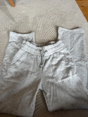 Grå mjukisbyxor med snörning - Säljer ett par ljusgrå mjukisbyxor med resår i midjan och snörning. Byxorna har två sidofickor och är tillverkade i mjukt bomullsmaterial. Perfekta för chill dagar eller när du vill vara extra bekväm.