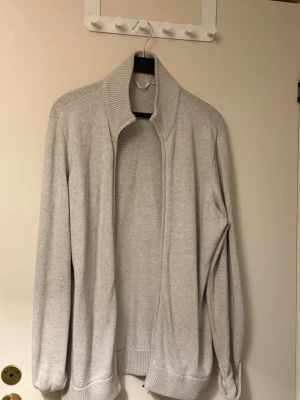 Ljusgrå ribbad kofta med krage - Ljusgrå kofta med ribbade muddar och hög krage. Koftan är öppen framtill utan knappar eller dragkedja och har långa ärmar. Perfekt för lager på lager och enkel att matcha med olika outfits.