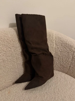 Bruna höga boots i mockaimitation - Säljer ett par bruna höga stövlar med spetsig tå och slouchy skaft. Stövlarna har dragkedja på insidan och är gjorda i mockaimitation. Perfekta för dig som vill ha en trendig och edgy look till hösten. Helt nya aldrig använda i kartongen 