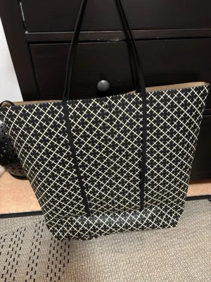  By Malene Birger Svart shoppingväska med vitt mönster - Stilren shoppingväska i svart med vitt rutmönster och långa svarta handtag. Väskan har en rymlig och fyrkantig form, perfekt för att bära mycket. Materialet ser ut att vara slitstark syntet eller konstläder. Passar dig som gillar grafiska mönster och enkel design. Populär väska som är perfekt till om man ska ta någon weekend❤️Höjd: 49 cm Bredd: 58 cm 