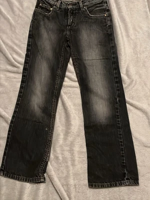 Svarta bootcut jeans med slitningar - Säljer ett par svarta bootcut jeans med snygga slitningar och tvättade detaljer. Jeansen har fem fickor, kontrastsömmar och klassisk knappgylf. Perfekta för en avslappnad och trendig look.