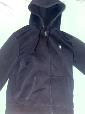 Svart hoodie från Polo Ralph Lauren - Svart hoodie med dragkedja från Polo Ralph Lauren. Klassisk design med vit broderad logga på bröstet, huva med dragsko och känguruficka framtill. Mjuk bomullsblandning som är skön att ha på sig.