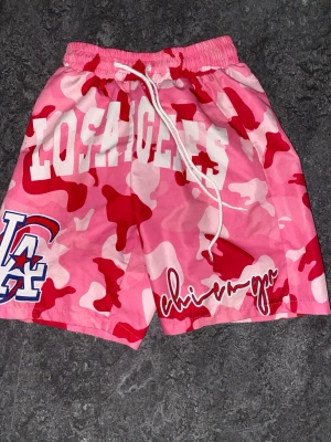Rosa camo shorts med LA-tryck - Färgstarka shorts med rosa, röd och vit camouflagemönster, stor 'Los Angeles'-text framtill och LA-logga. Justerbar midja med snörning och bakficka. Perfekta för dig som vill sticka ut i sommar.