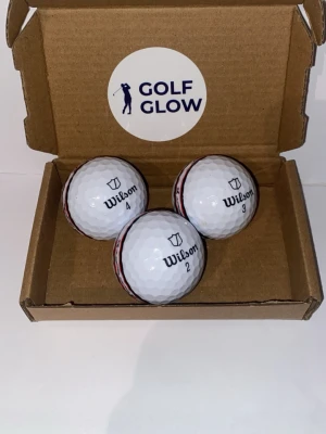 Wilson duosoft stripe 3-pack  - Tre vita golfbollar från Wilson med svart och röd detalj, levereras i en brun presentbox med Golf Glow-logga. Perfekt för golfentusiasten som vill ha kvalitetsbollar med snygg design.