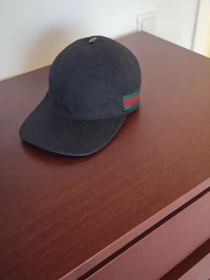 Svart Gucci keps med GG-mönster - Säljer en svart keps från Gucci med klassiskt GG-mönster över hela kepsen och den ikoniska gröna och röda randen på sidan. Justerbar rem baktill för perfekt passform. Snygg och stilren accessoar för dig som gillar exklusiva märken.