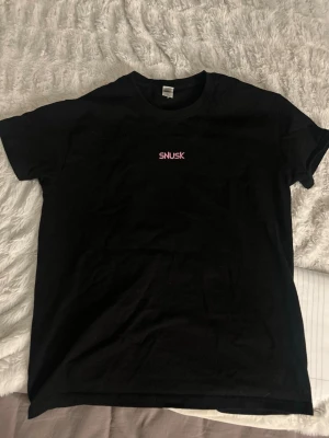 Svart SNUSK t-shirt med rosa tryck - Svart t-shirt från Fröken Snusk med rosa text på bröstet och en stor rosa siluett av en ryttare på häst på ryggen. Använd fåtal gånger!