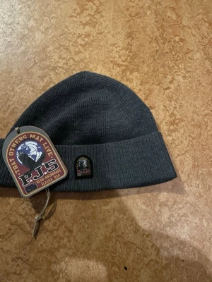 Mörkgrå mössa från Parajumpers - Snygg mörkgrå ribbstickad mössa från Parajumpers med uppvikt kant och klassisk PJS-logga framtill. Perfekt för kalla dagar och enkel att matcha med olika outfits. Tillverkad i mjukt material som sitter skönt på huvudet.