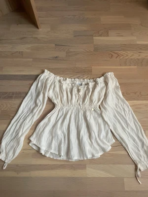 Offshoulder cremevit blus med resår - Trendig offshoulder blus i cremevitt med långa puffiga ärmar och resår i midjan och vid axlarna. Tillverkad i ett lätt, luftigt tyg. Perfekt för att styla med jeans eller kjol. Superfin inför sommaren!