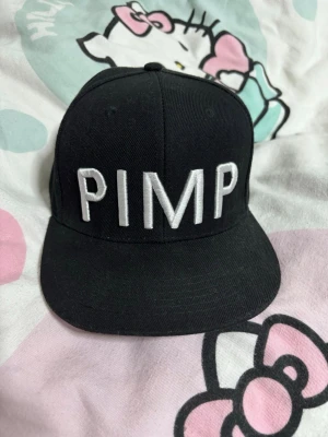 Svart snapback keps med PIMP-tryck - Cool svart snapback keps med stort vitt broderat PIMP-tryck på framsidan. Klassisk platt skärm och justerbar baksida med plastspänne. Perfekt för dig som vill sticka ut och addera attitude till din outfit.