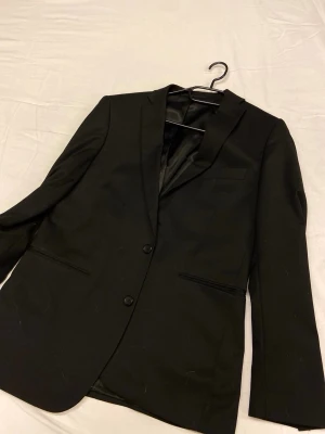 Dressmann kostym  slim fit - Stilren svart kavaj från Dressmann i slim fit-modell. Klassisk design med två knappar framtill, smala slag och innerfoder i svart. Passar perfekt till både skjorta och t-shirt för en snygg look.