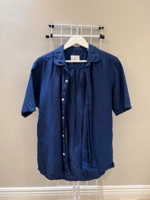 Blå kortärmad linneskjorta - Snygg blå skjorta i linne med korta ärmar och klassisk krage. Skjortan har knappar framtill och en avslappnad passform som ger en chill vibe. Perfekt för varma dagar och enkel att styla med jeans eller shorts.