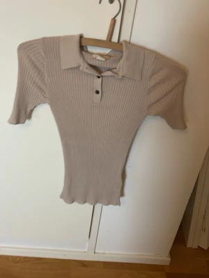 Beige ribbad polotopp med knappar - Säljer en beige ribbad polotopp med korta ärmar och klassisk krage. Toppen har knappar framtill och är figurnära i modellen. Perfekt till jeans eller kjol för en clean och stilren look.