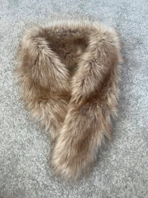 Beige fuskpäls halsduk - Säljer en fluffig beige fuskpäls halsduk med mjukt satinliknande foder på insidan. Halsduken har en smart ögla för att hålla den på plats och ger en lyxig känsla till din outfit, spännet är trasigt med går fortfarande att använda . Perfekt för dig som vill ha något extra mysigt och trendigt.