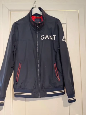 Marinblå vindjacka från GANT - Snygg marinblå vindjacka från GANT med vita logotryck på bröstet och ryggen. Jackan har dragkedja framtill, två fickor med röda dragkedjor och ribbade muddar med vita ränder vid ärmslut och nederkant. Insidan av kragen har röd detalj. Perfekt för en sportig look.