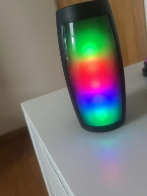 Bärbar svart högtalare med LED-ljus - Snygg bärbar högtalare i svart plast med coolt LED-ljus som skiftar i flera färger. Högtalaren har cylinderformad design, högtalaren på baksidan och flera anslutningsmöjligheter på toppen. Perfekt för att ta med sig och spela musik trådlöst. Kopplas med Bluetooth 😊