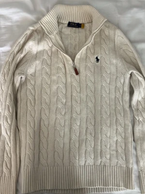 Vit kabelstickad tröja Polo Ralph Lauren - Kabelstickad vit tröja från Polo Ralph Lauren med halv dragkedja och ribbad krage. Tröjan har det klassiska mörkblå logobroderiet på bröstet och ribbade muddar. Perfekt för en clean och stilren look.