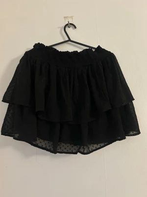 Svart volangkjol med prickigt tyg - Säljer en svart kortkjol med dubbla volanger och transparent prickigt tyg i understa lagret. Kjolen har resår i midjan och en luftig, flowy känsla som passar perfekt till sommaren. Enkel att matcha med olika toppar för en trendig look.