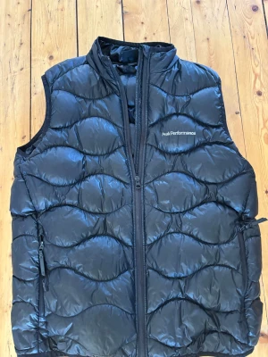 Svart dunväst Peak Performance - Snygg svart dunväst från Peak Performance med vågformat quiltat mönster. Västen har hög krage, två dragkedjeförsedda fickor och Peak Performance-logga på bröstet. Tillverkad i glansigt material och fylld med dun för extra värme.