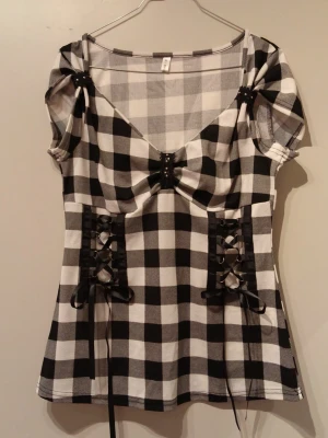 Rutig topp med snörning  - Säljer en svartvit rutig topp med korta puffärmar och snygg snörning i midjan. Toppen har en v-ringad urringning med liten rosett och är tillverkad i mjuk polyester. Perfekt för dig som gillar statement-plagg med coola detaljer.