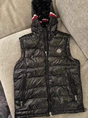 Svart dunväst från Moncler med huva - Snygg svart dunväst från Moncler med glansig finish, två dragkedjefickor och huva med röd och vit 