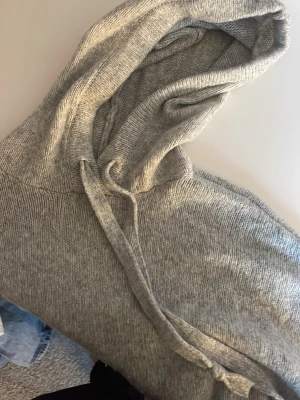 Grå stickad hoodie med dragsko - Mysig grå stickad hoodie med huva och dragsko. Tröjan har långa ärmar och är tillverkad i ett mjukt, melerat material som känns skönt mot huden. Perfekt för dig som gillar en enkel och stilren look.
