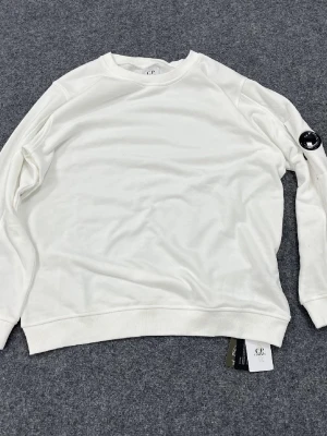 Vit sweatshirt från C.P. Company - Vit sweatshirt från C.P. Company med rund halsringning och ribbade muddar. Tröjan har en ikonisk svart linsdetalj på ena ärmen och är tillverkad i mjukt bomullsmaterial. Perfekt för en clean och stilren look.