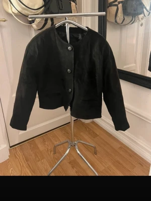 Svart mocka bomberjacka - Snygg svart bomberjacka i mocka med rund halsringning och knäppning framtill. Jackan har en croppad passform och två fickor fram. Perfekt för dig som vill ha en stilren och trendig look.