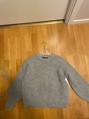 Grå stickad tröja från BikBok - En enkel och stilren grå stickad tröja med rund halsringning och ribbade muddar vid ärmslut och nederkant. Tröjan har en avslappnad passform och är perfekt för lager på lager under kyliga dagar. Materialet ser ut att vara mjukt och bekvämt. Vid halsringningen finns små få rosa prickar kan kanske gå bort vid tvätt vet ej