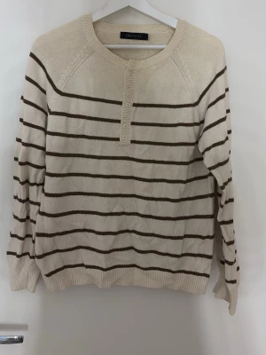Beige stickad tröja med bruna ränder - Snygg beige stickad tröja från Costume med bruna horisontella ränder. Tröjan har rund halsringning, knappslå framtill och långa ärmar. Ribbstickade muddar vid ärmslut och nederkant. Perfekt för en avslappnad och trendig look. Köpt i Frankrike