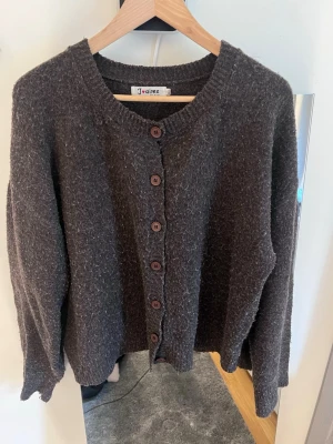 Mörkbrun kofta från J'aime Olyto - Mysig mörkbrun kofta från J'aime Olyto med rund halsringning och knappar framtill. Koftan är stickad med en lite luddig yta och har långa ärmar samt ribbade muddar vid hals och ärmslut. Perfekt för lager på lager och chill stil.